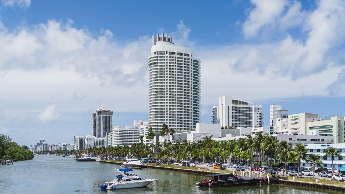 Miami Beach waterfront with a curved luxury condo tower, marina, and palm-lined promenade