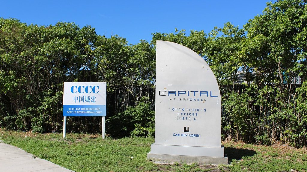 A weathered monument sign reading Capital at Brickell, condominiums, offices and retail, standing in front of an overgrown empty lot